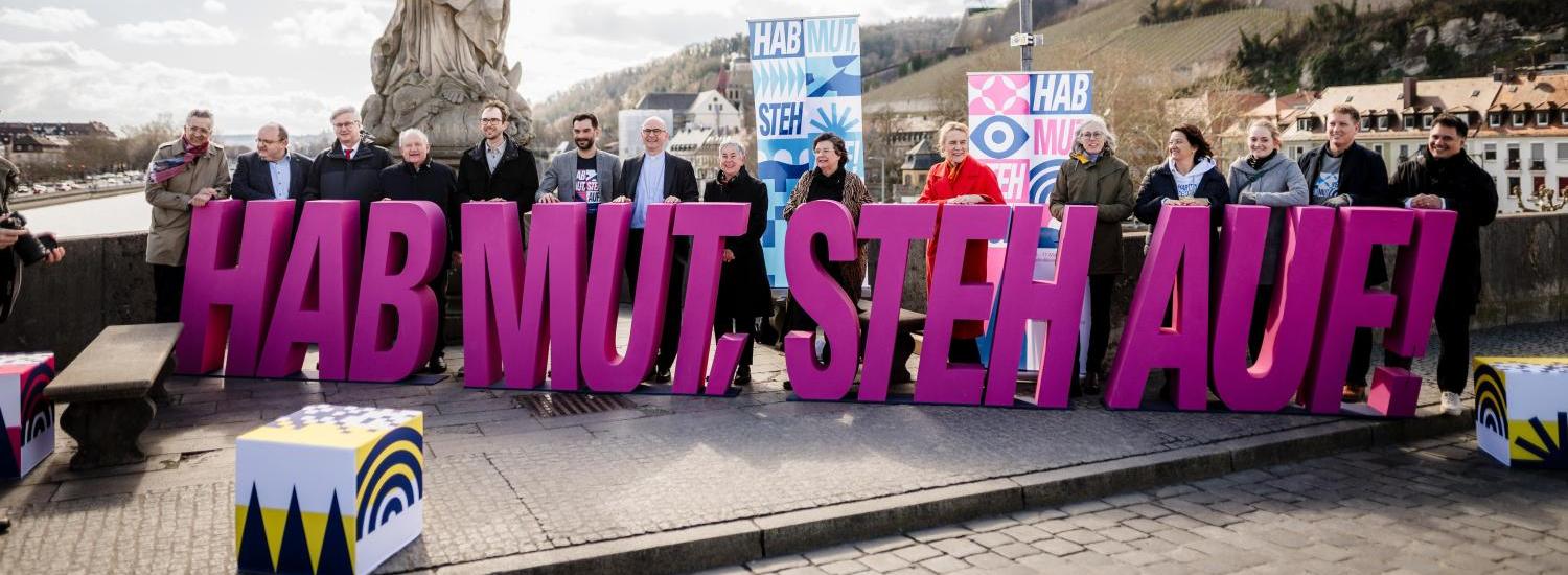 Menschen halten auf Brücke in Würzburg große, pinke Buchstaben, die den Schriftzug „HAB MUT. STEH AUF!“ bilden.