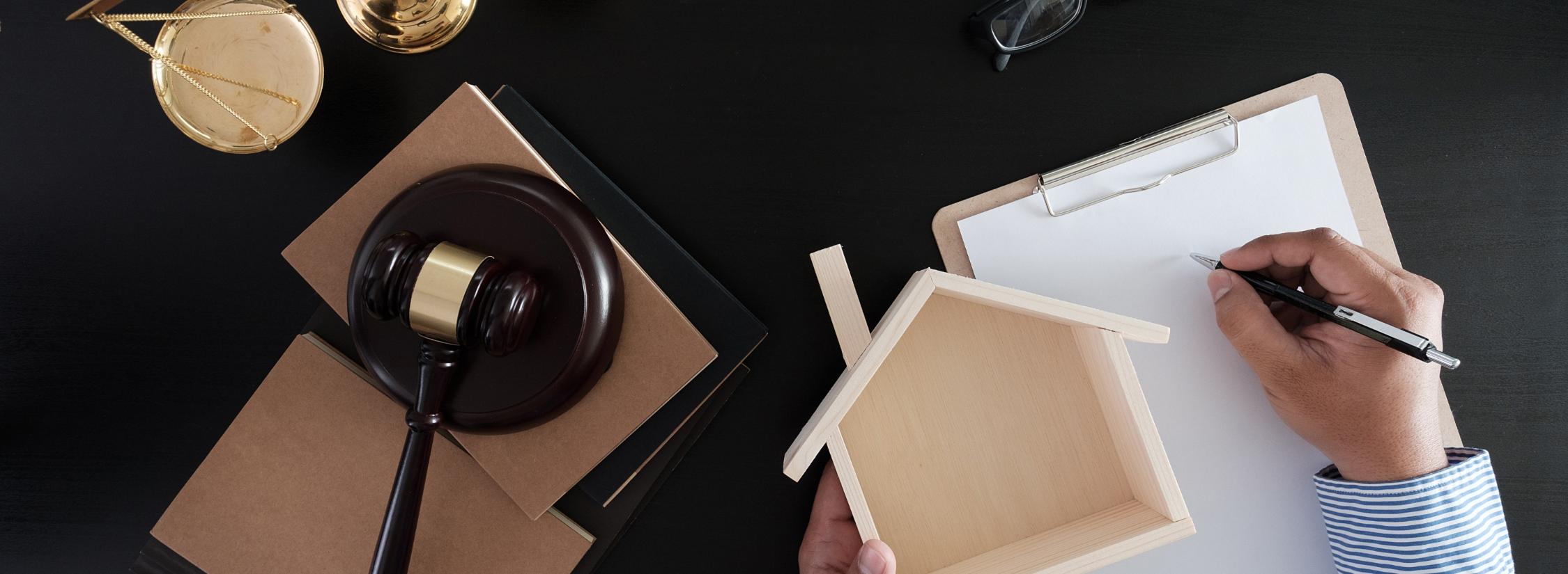 Erben und Vererben Jemand hält ein Modell eines Hauses in der Hand und schreibt etwas auf. Daneben sehen eine Waage und ein Richterhammer, die auf Justitia hinweisen.