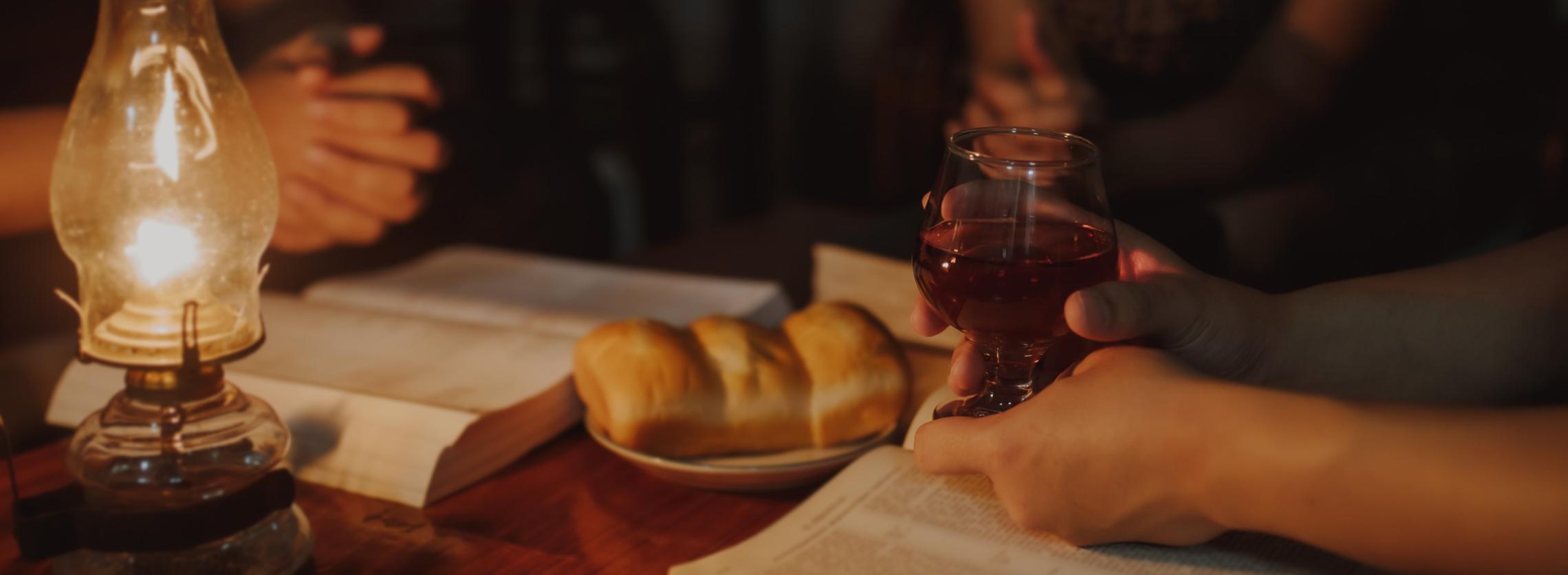 Christliche Familie feiert zu Hause Eucharistie mit Brot und Wein. Auf dem Holztisch steht zudem eine Öllampe und es liegen dort zwei aufgeschlagene Bücher