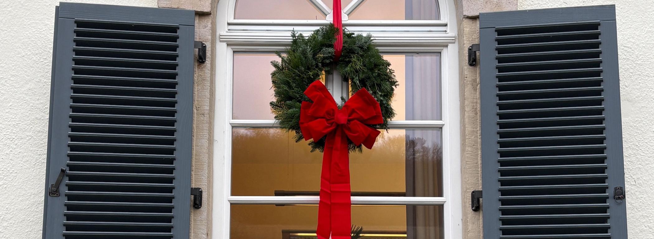 Türkranz mit Tannenzweigen und roter Schleife von außen an einem Rundbogenfenster im oberen Stockwerk mit geöffneten Fensterläden