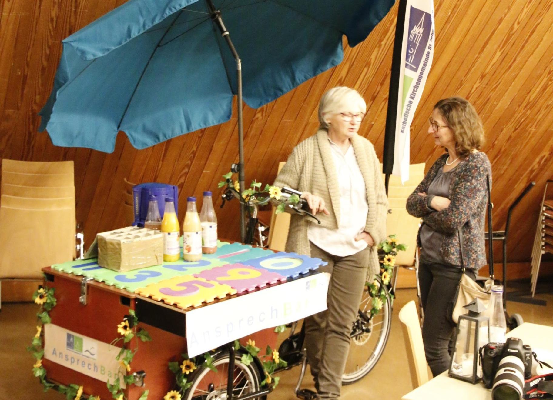 Lastenfahrrad, das als AnsprechBar aufgestellt werden kann, mit Getränken, Sonnenschirm und Beachflag mit Gemeindelogo ausgestattet und 2 Personen, die sich unterhalten