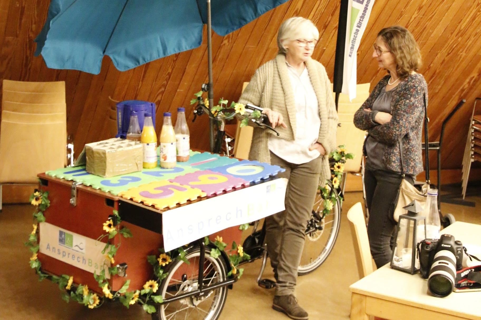 Lastenfahrrad, das als AnsprechBar aufgestellt werden kann, mit Getränken, Sonnenschirm und Beachflag mit Gemeindelogo ausgestattet und 2 Personen, die sich unterhalten
