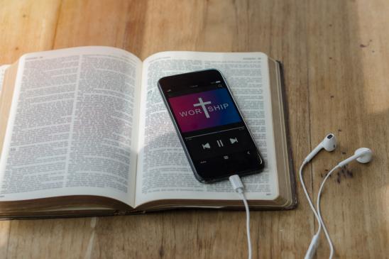 Aufgeschlagene Bibel mit Smartphone mit Warship-App und Ohrstöpsel auf einem Holzuntergrund