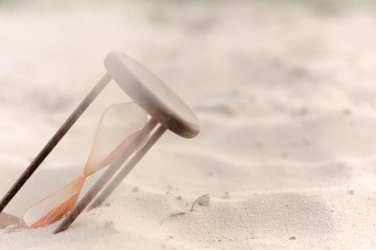 Sanduhr, die schräg im Sand steckt, was den Fluss des Sandes verlangsamt