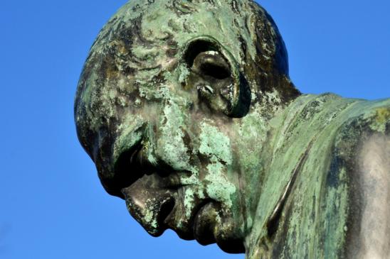 Kopf einer Bronzestatue mit viel Patina. Der Kopf ist geneigt, der Blick geht nach unten.